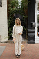 A woman with wavy blonde hair sits on a wooden bench holding a small iced coffee in one hand and a fluffy Pomeranian dog in the other. She is wearing light-colored, coffee cup-patterned pants and studded black boots