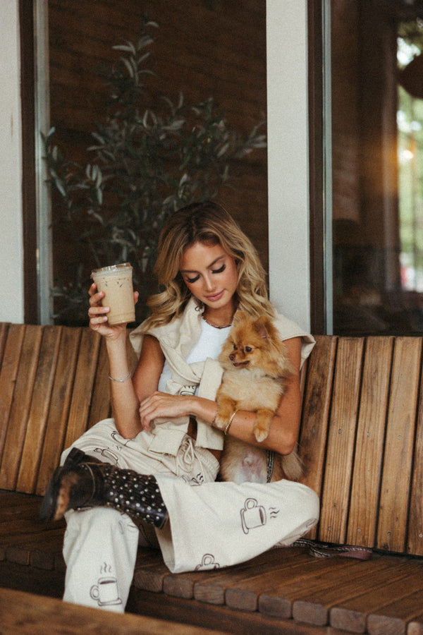 A stylish woman sits relaxed on a bench with a Pomeranian on her lap, sipping iced coffee. She wears light beige pants with coffee cup prints and a casual sleeveless top